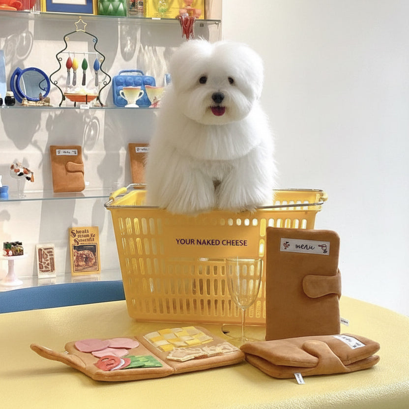 Small white dog in a yellow basket with cheese-themed items on a table.